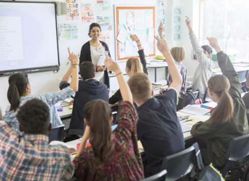 A classroom with teenage pupils raising their hands, a teacher in front of them