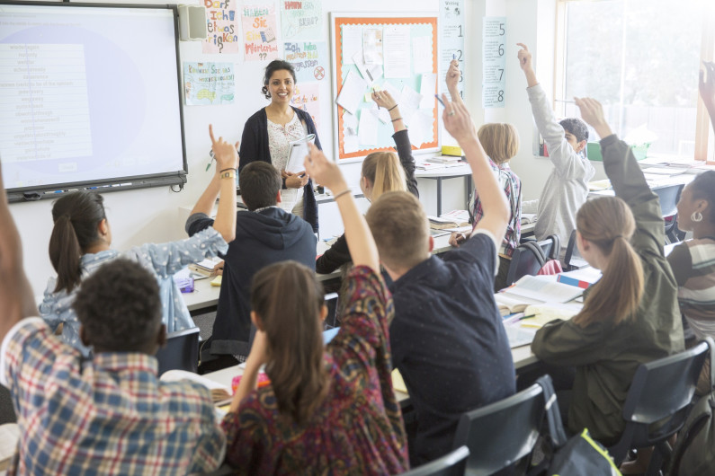 A classroom with teenage pupils raising their hands, a teacher in front of them